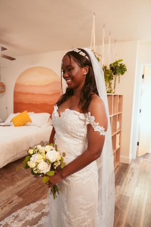 Bride in a white lace wedding dress with off-shoulder details holds a cream and white bouquet in a bedroom setting.