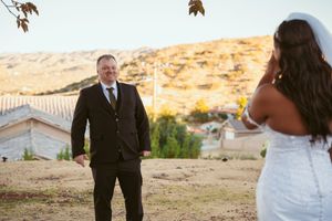 Bride in flowing white dress and groom in dark suit share a moment on hillside during wedding.