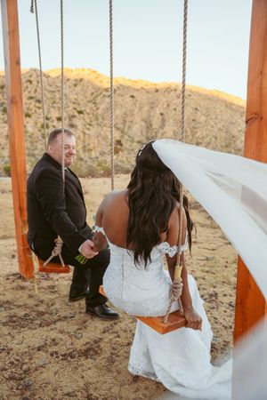 Couple shares playful moment on wooden swing set against desert mountain backdrop.