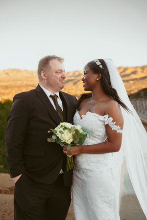 Wedding couple shares intimate moment on desert hillside while holding white bouquet at golden hour.