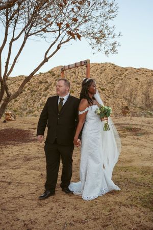 A couple in wedding attire stands together in a desert landscape with mountains and bare trees in the background.