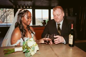 A wedding couple shares an intimate toast together at a dark wooden table with wine.