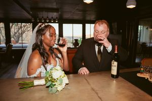 Newlywed couple enjoys drinks at bar with white floral bouquet and wine bottle on counter.