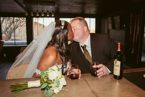 Newlywed couple shares intimate moment at a wooden bar table with wine and bridal bouquet.