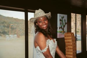 A joyful figure in a white beaded dress and sparkly cowboy hat smiles while posing indoors near a window.