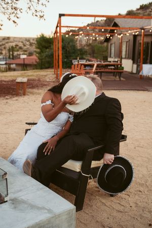 A couple in wedding attire share a romantic moment under a rustic wooden pergola with string lights.