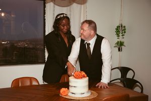 A couple stands over their white wedding cake topped with fresh orange flowers at an intimate indoor reception.