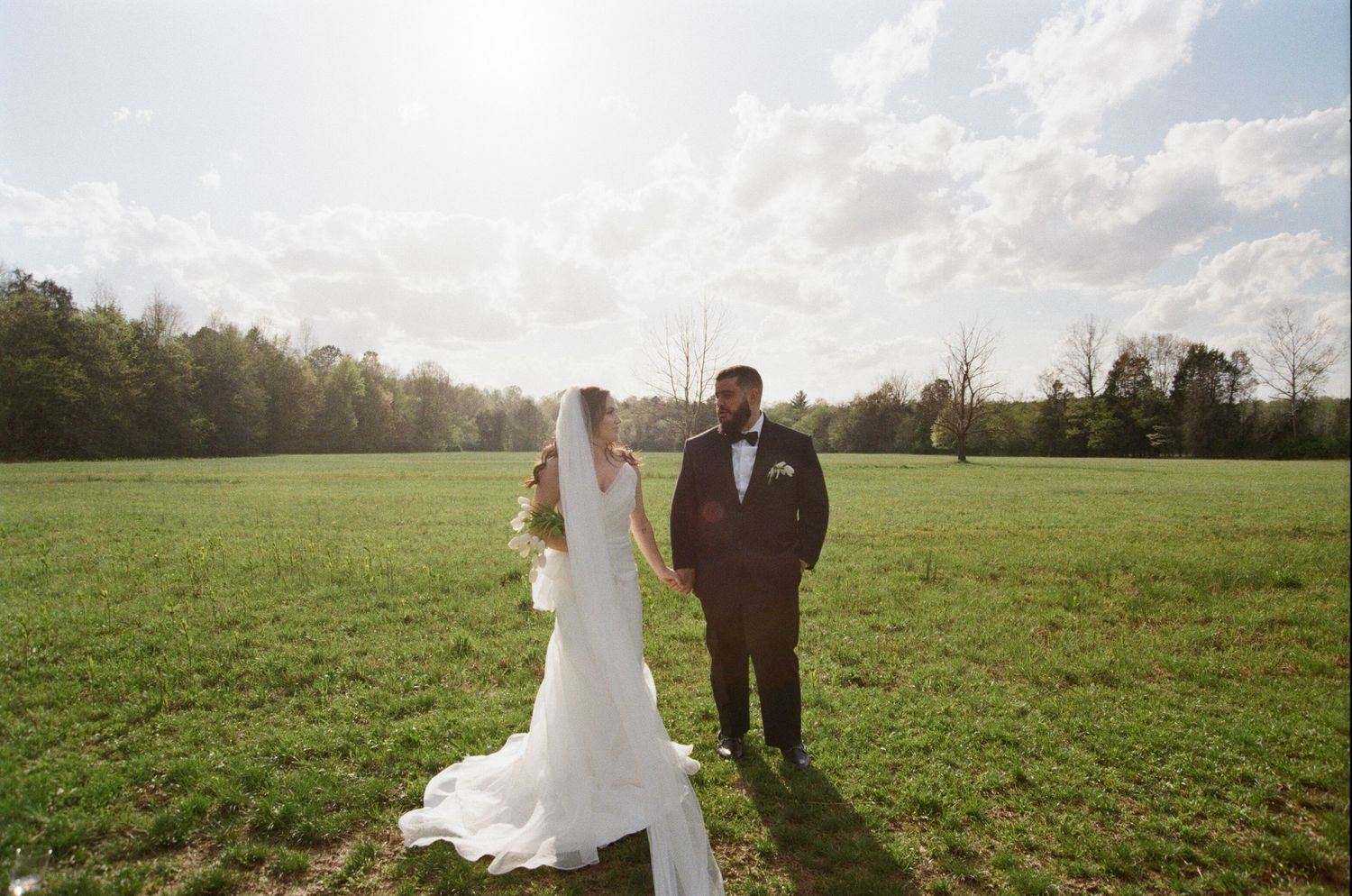A wedding couple stands together in a sun-lit green field with trees in the background on a beautiful spring day.