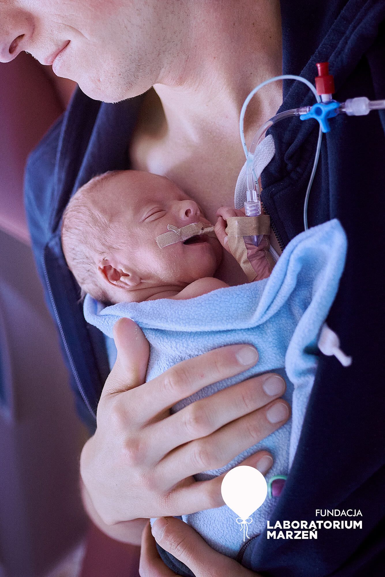 Ojciec trzymający w ramionach wcześniaka na oddziale neonatologii, zdjęcie z wystawy Fundacji Laboratorium Marzeń – autorstwa Anety Ludwig.