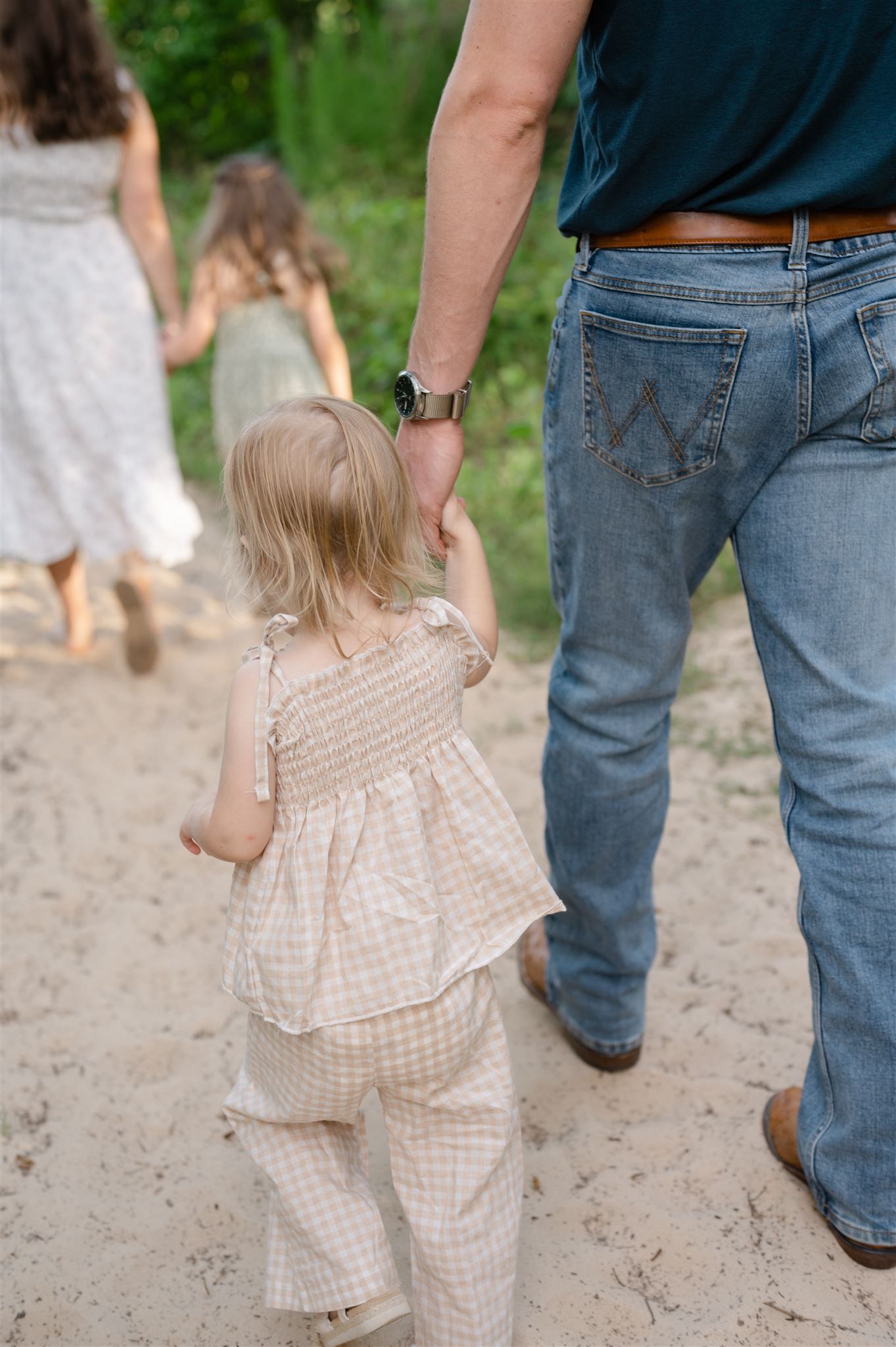 A small child in checkered outfit holds hands with parents while walking on a dirt path in nature.