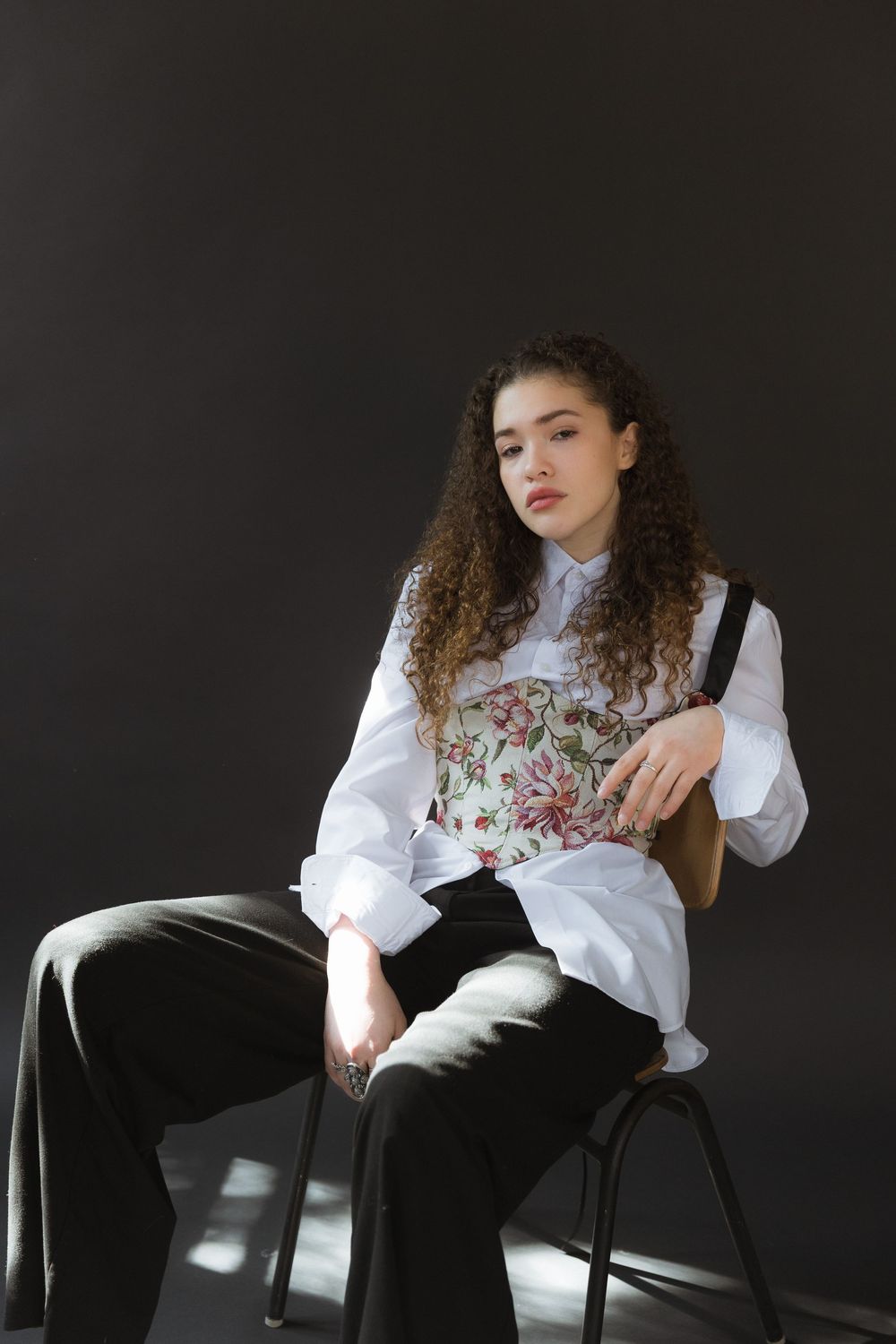 A model in a white blouse and floral corset sits on a chair against a dark backdrop in dramatic lighting.