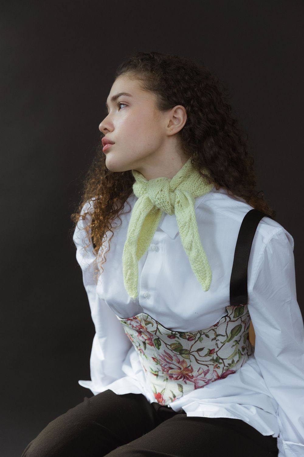 Portrait of someone in profile wearing a white button-up shirt, lime green neck scarf and floral corset against dark background.
