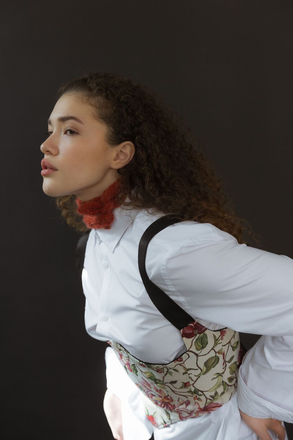 A profile view against dark background showing curly hair, white blouse, red scarf and floral print corset.
