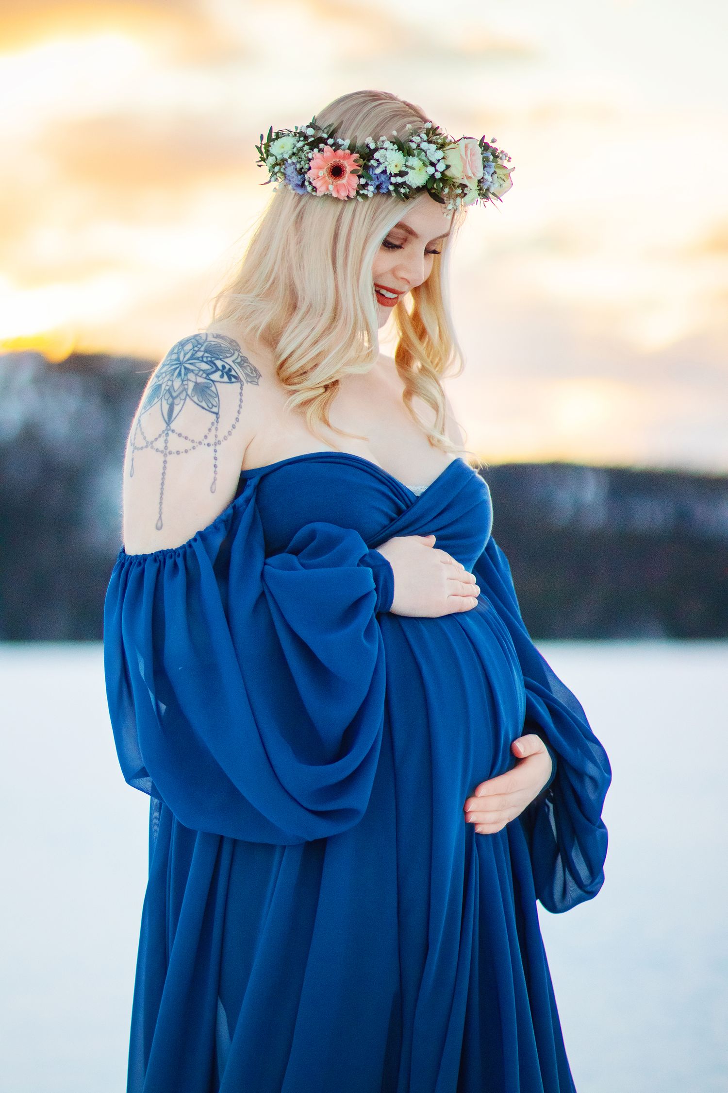 Pregnant blonde woman in blue off-shoulder gown with floral crown at winter sunset landscape.