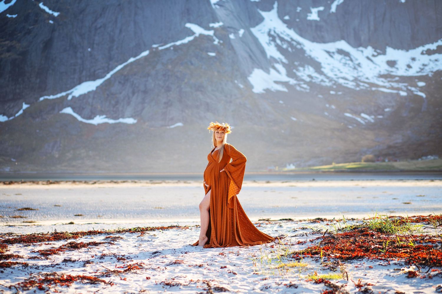 Woman in rust orange gown with floral crown on snowy beach with mountain backdrop.