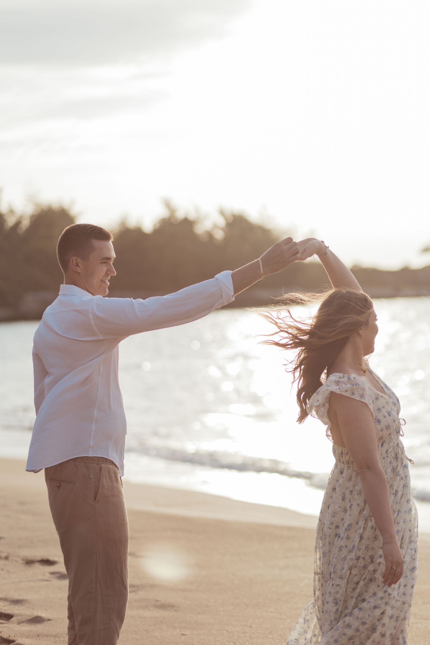 A couple shares a playful moment dancing together at sunset on a sandy beach with ocean waves in the background.