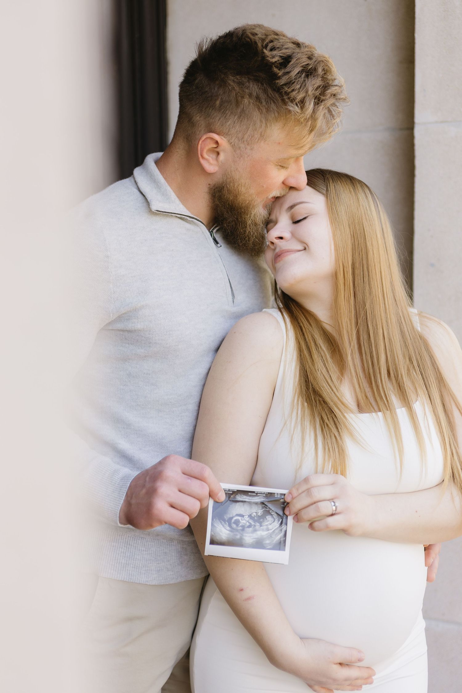 Couple shares intimate moment while holding ultrasound photo against wall.