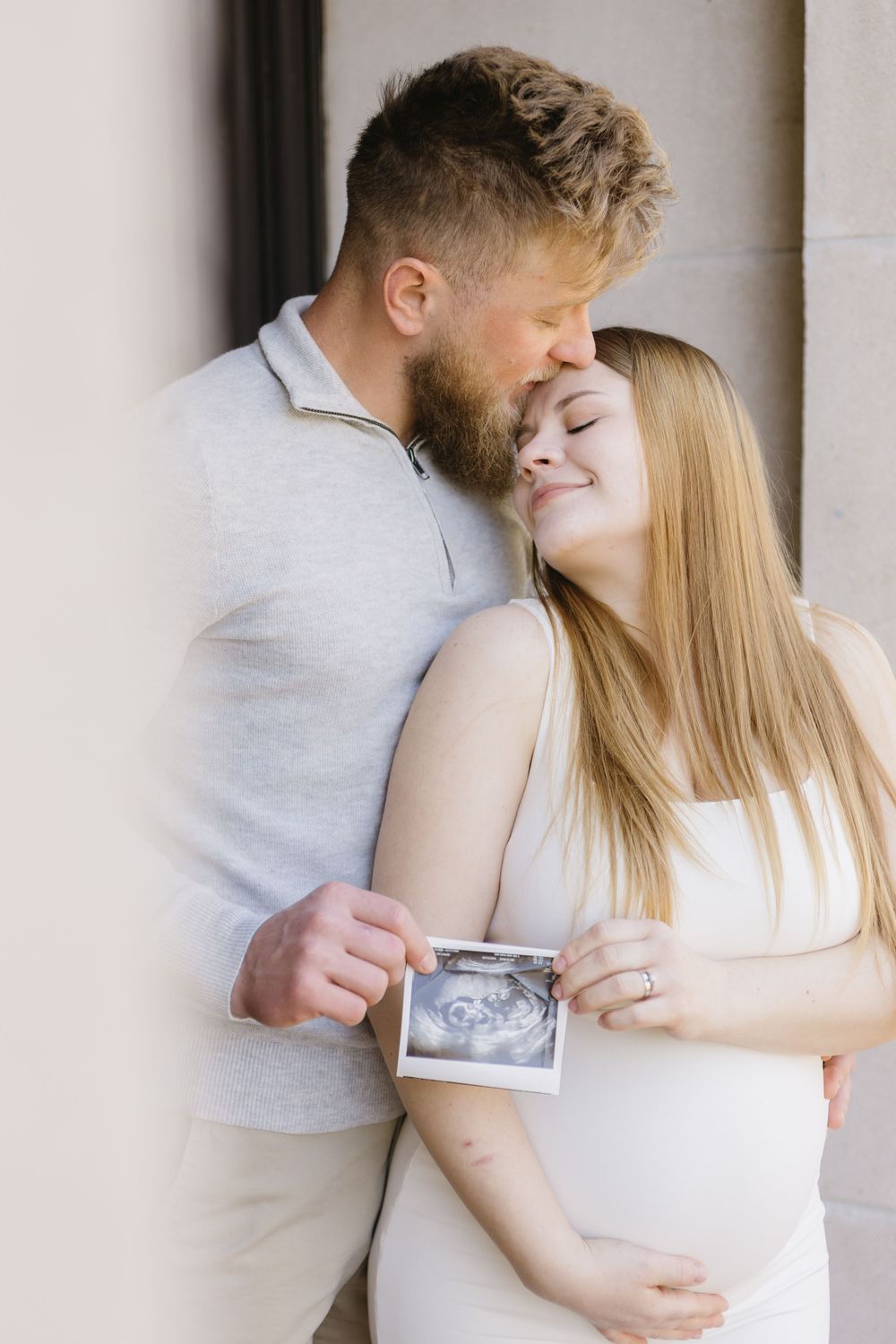 Couple shares intimate moment while holding ultrasound photo against wall.