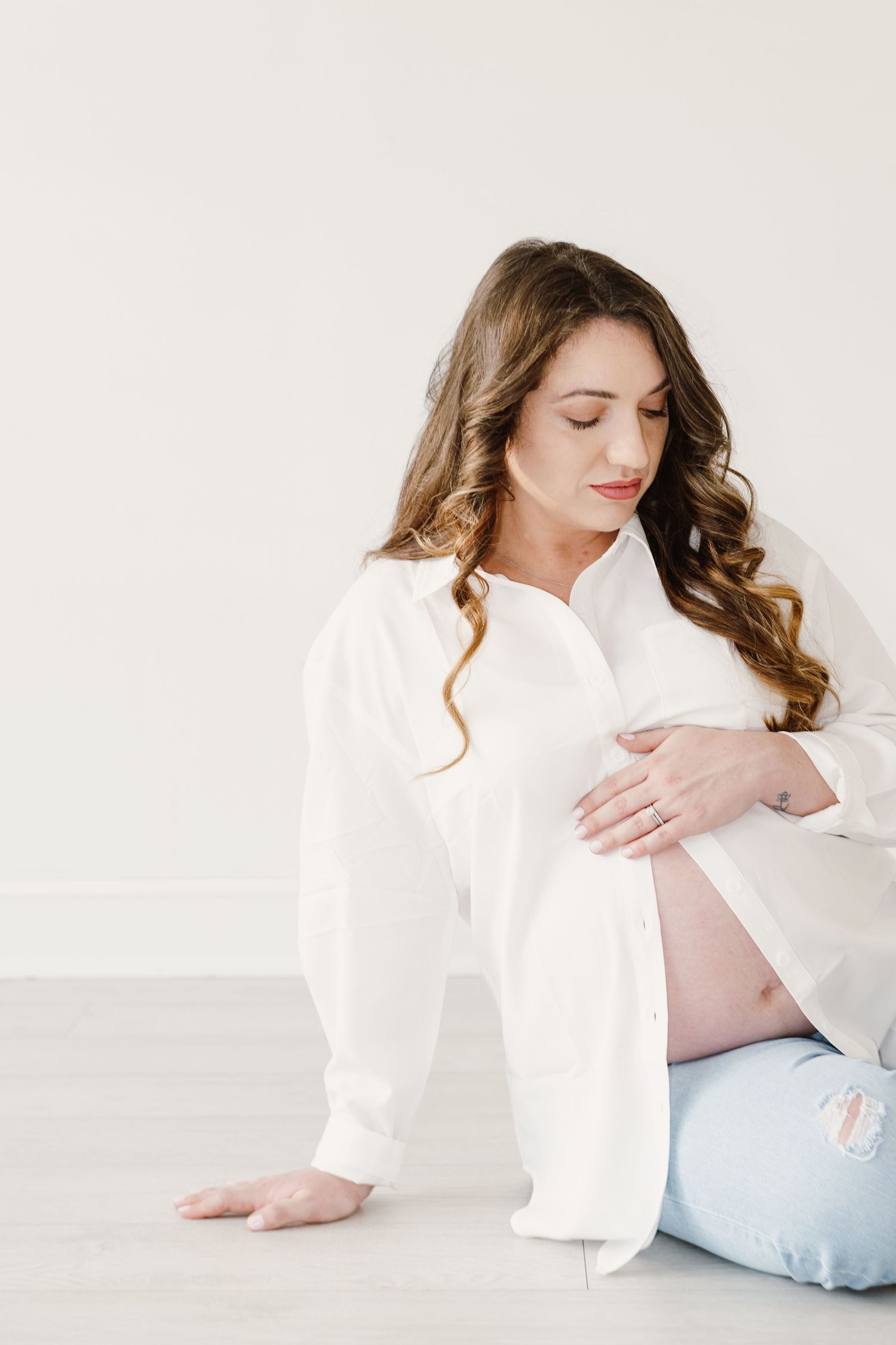 Expectant woman in white button-down shirt sits peacefully against light background.