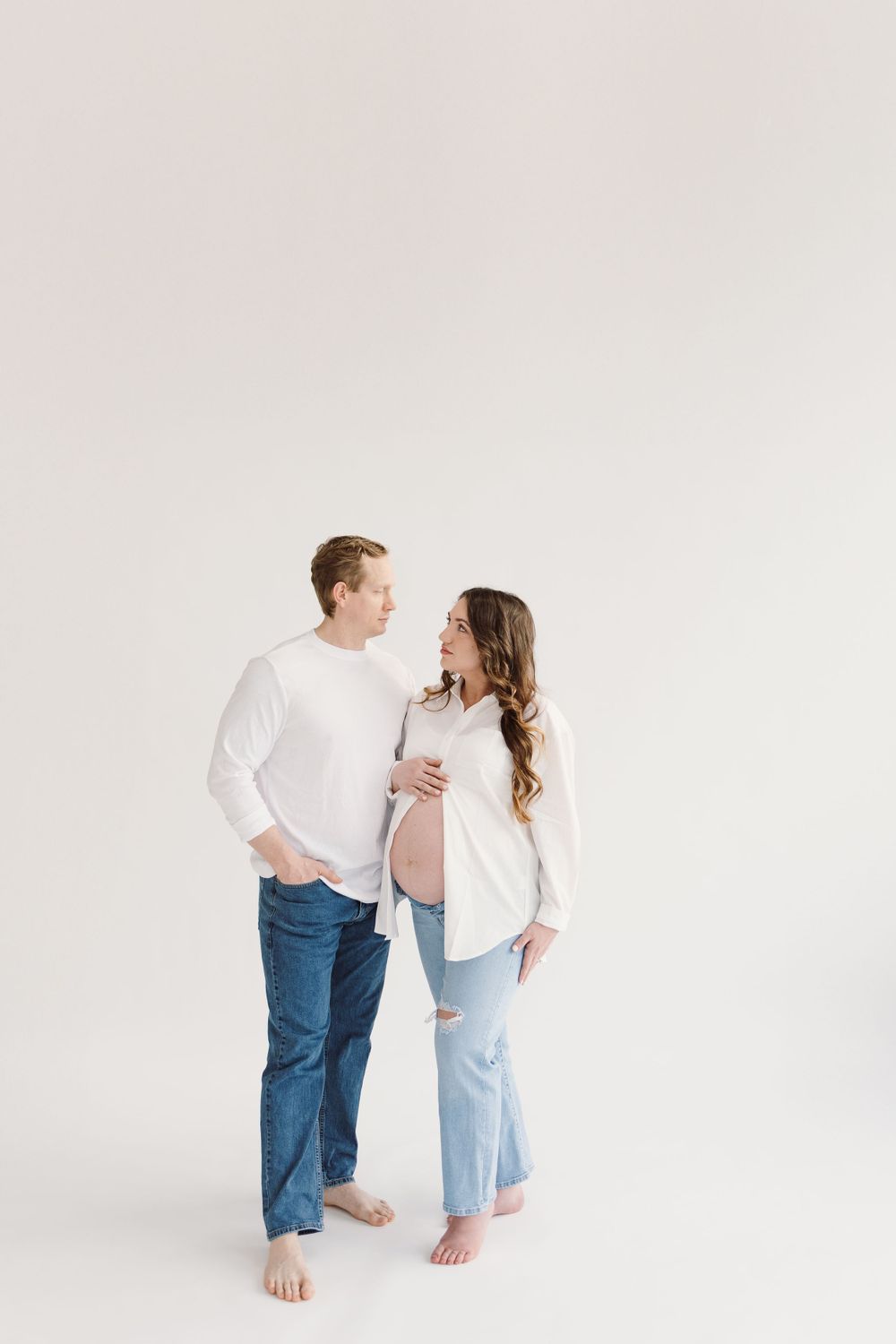 Barefoot couple in white tops and jeans share tender moment against white wall.
