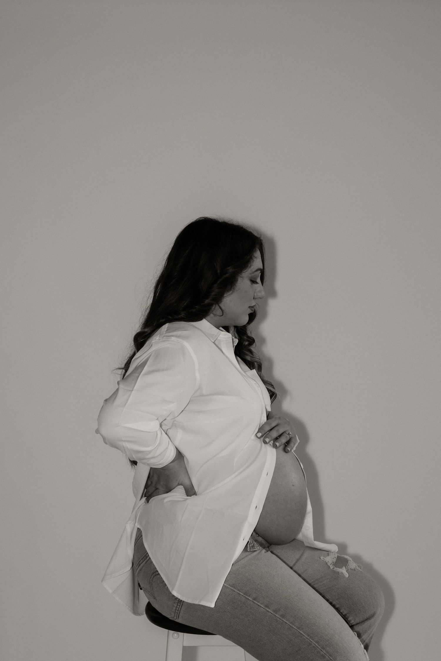 Side profile of pregnant woman in white shirt and jeans against neutral backdrop.