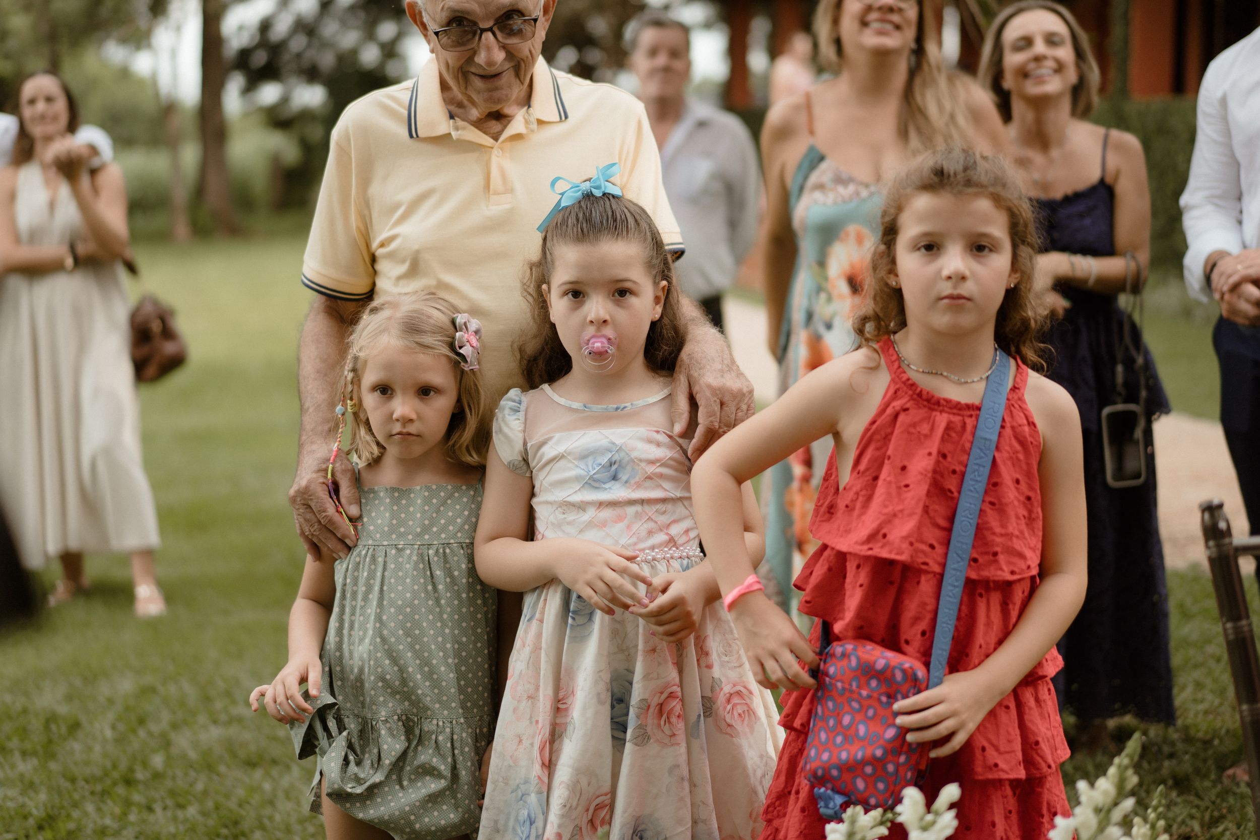 Three young children in summer dresses watching the wedding festivities on a grassy lawn.