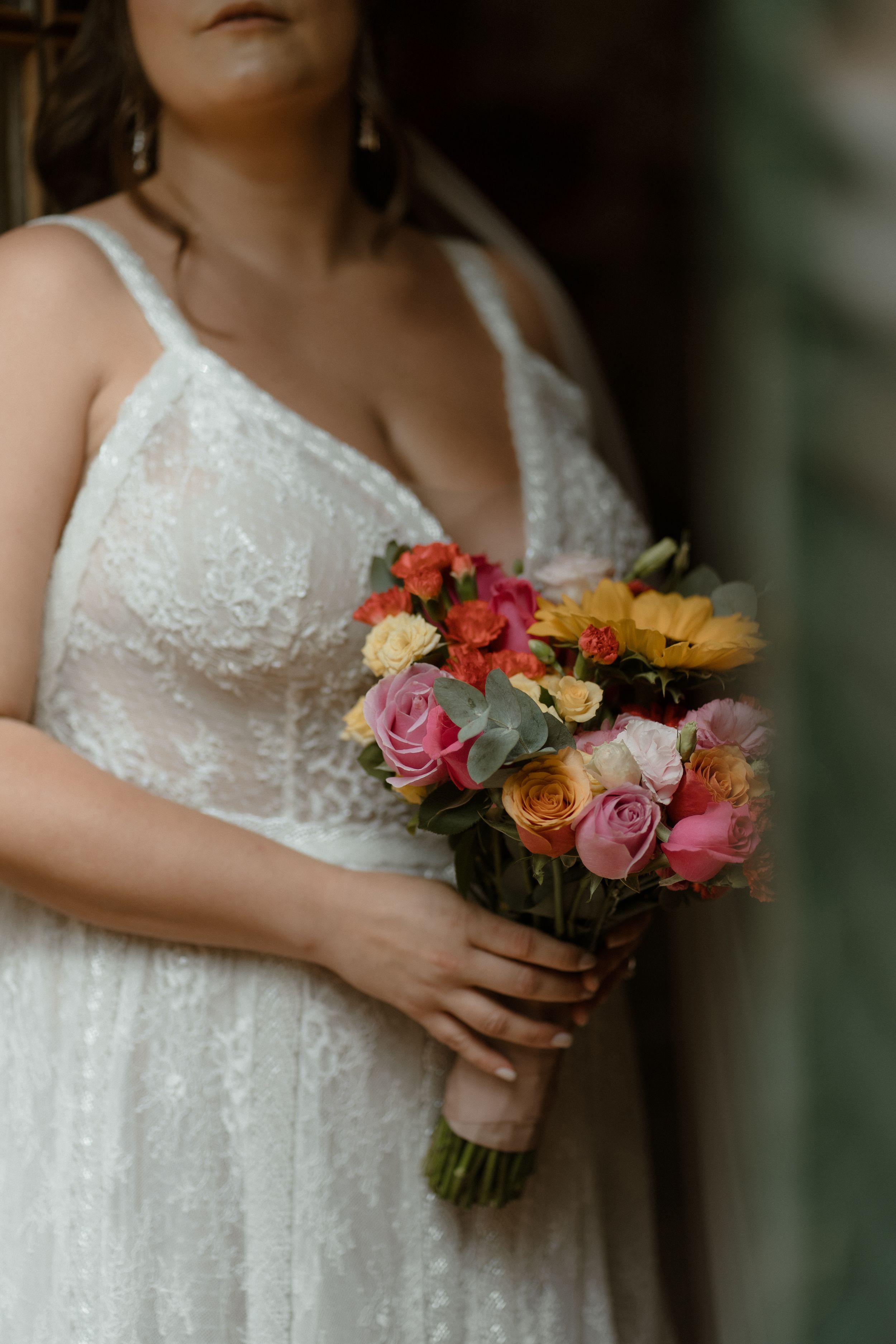 Close-up of colorful wedding bouquet with pink, orange and yellow roses held by person in white lace dress.