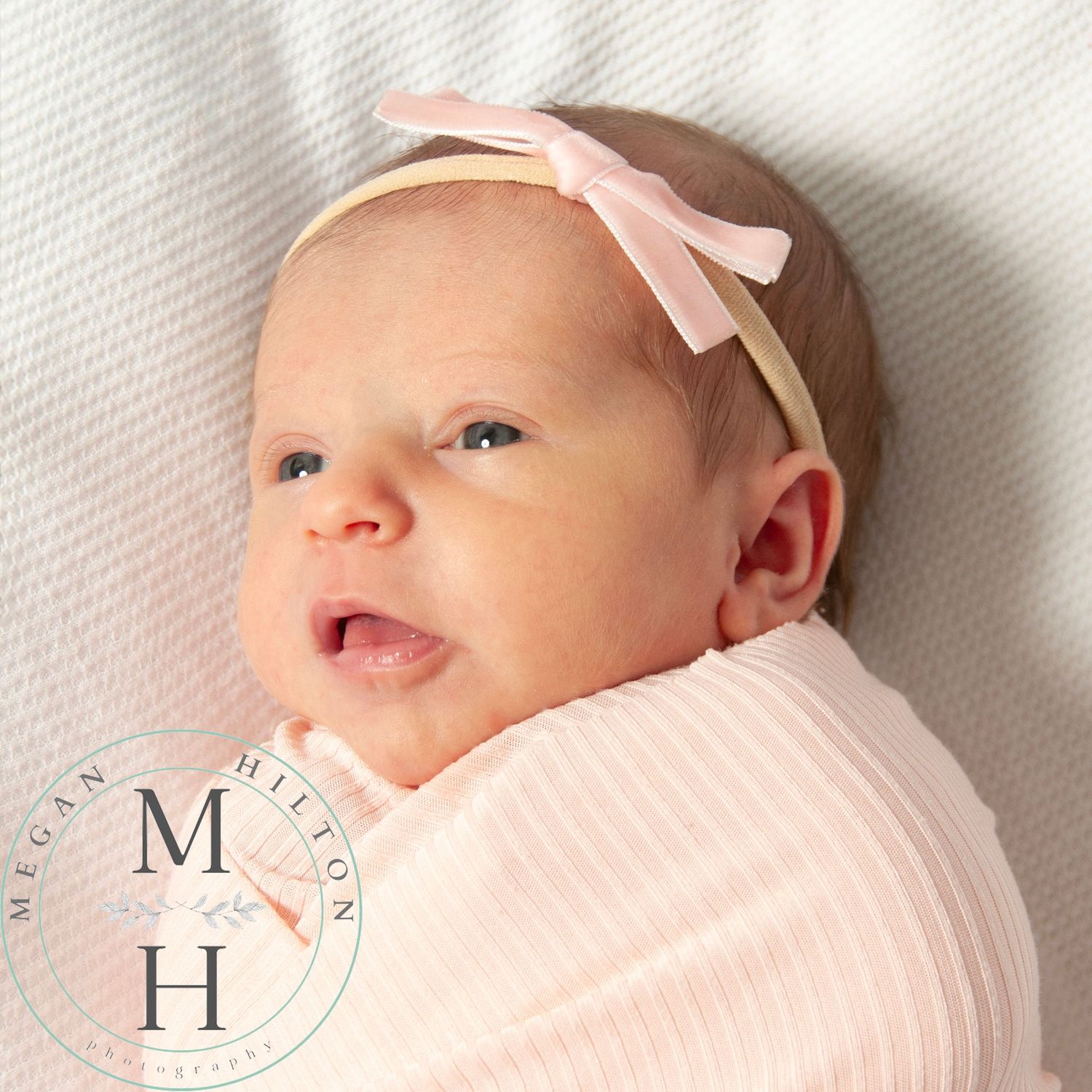 A tiny baby swaddled in soft pink fabric wearing a light pink bow headband, photographed against a white background.