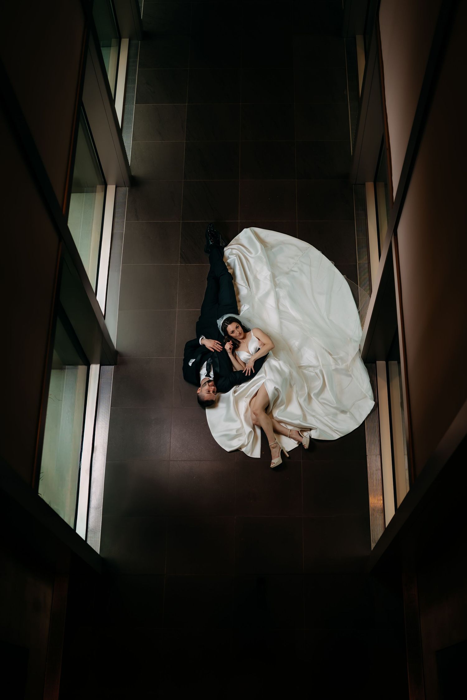 High-angle view of newlyweds in an elegant dress and suit lying on dark floor in modern hallway Grand Rapids Art Museum Michigan