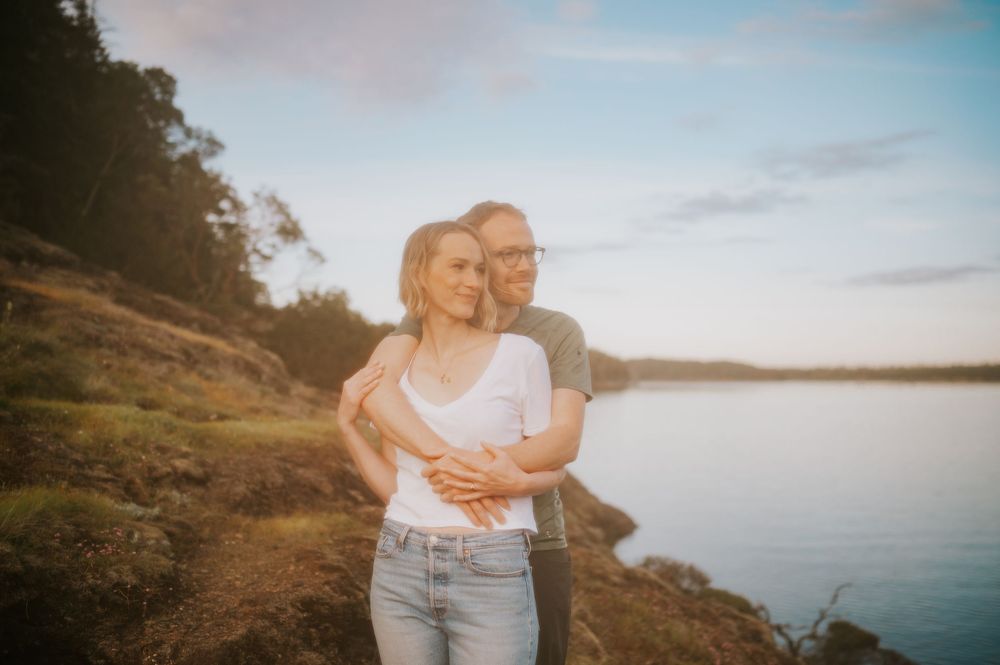 Engaged couple standing on rocky cliff overlooking Comox Lake, hugging during sunset for couple adventure session photoshoot