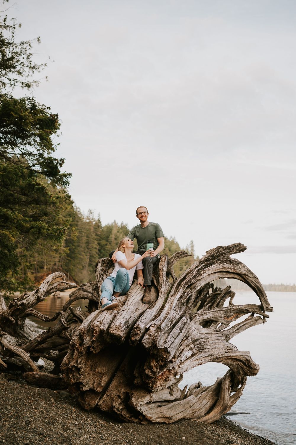 Couple sitting on tree stump by Comox Lake with water bottle during photography session, the couple’s engagement photoshoot