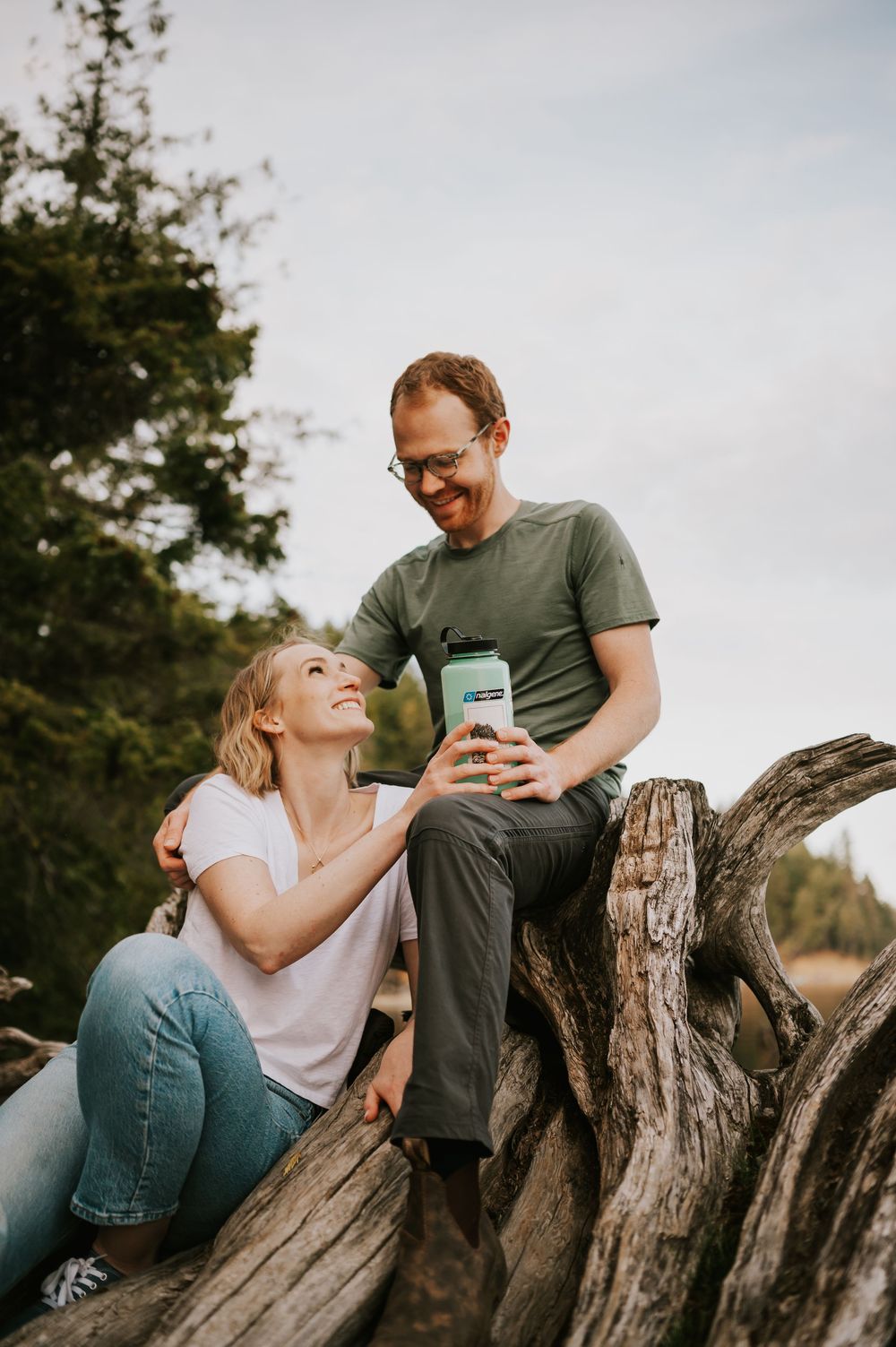 Couple sitting on tree stump by Comox Lake laughing and holding water bottle during photography session by soulful adventure photographer