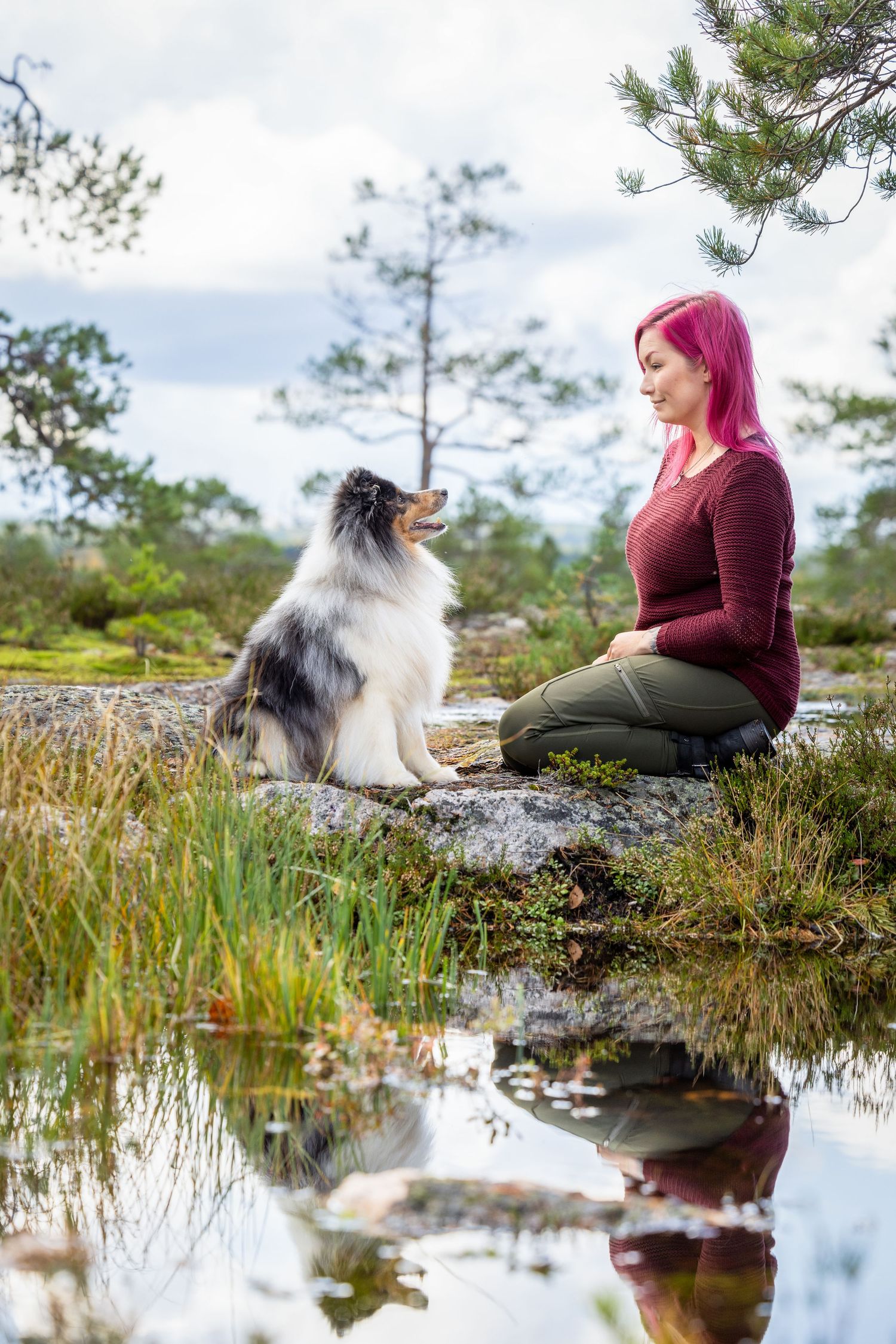 A Shetland Sheepdog sits with a person in burgundy sweater by a tranquil pond reflecting trees and sky.
