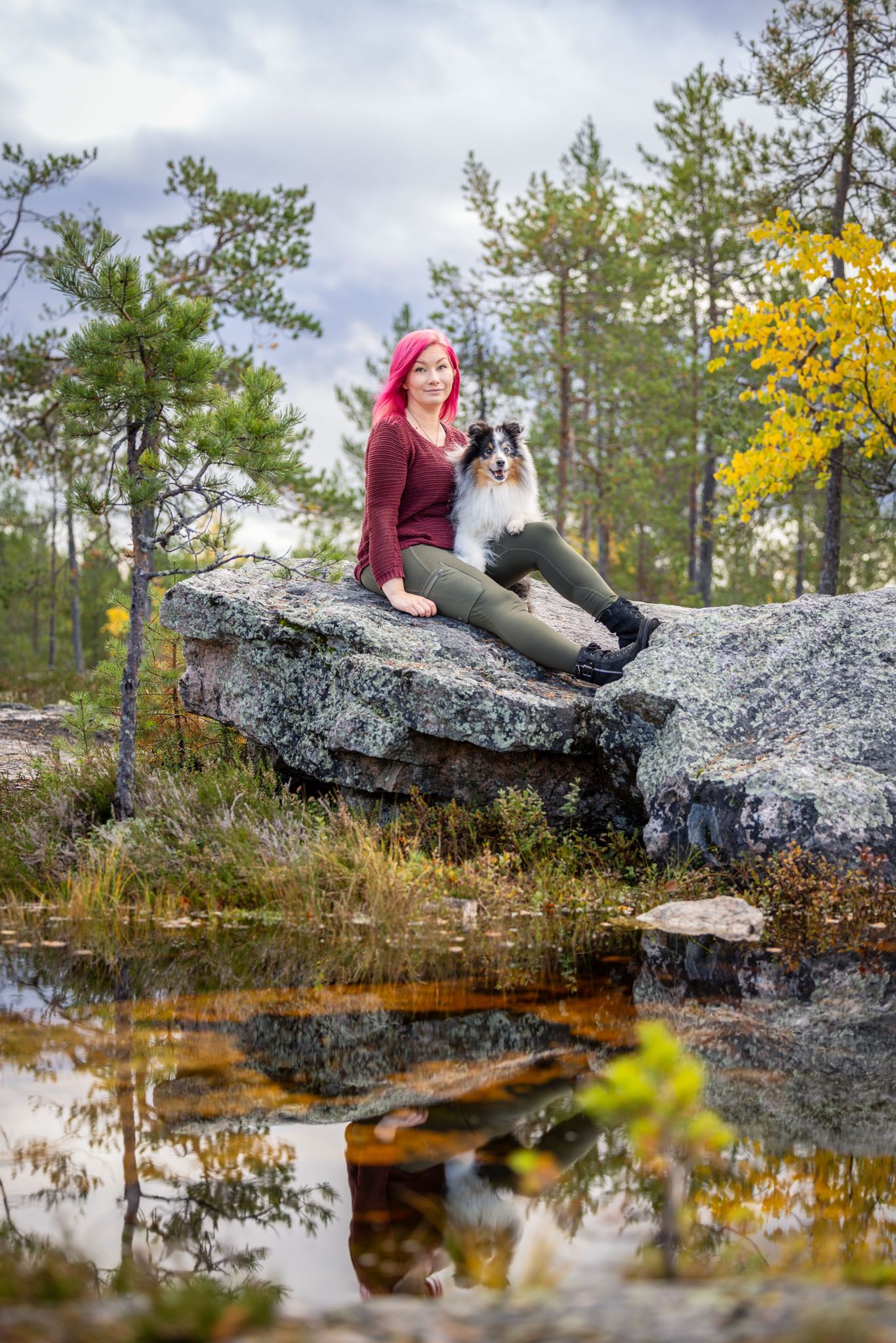 A border collie and someone sit on a large rock formation near a reflective pond surrounded by pine trees and autumn foliage.