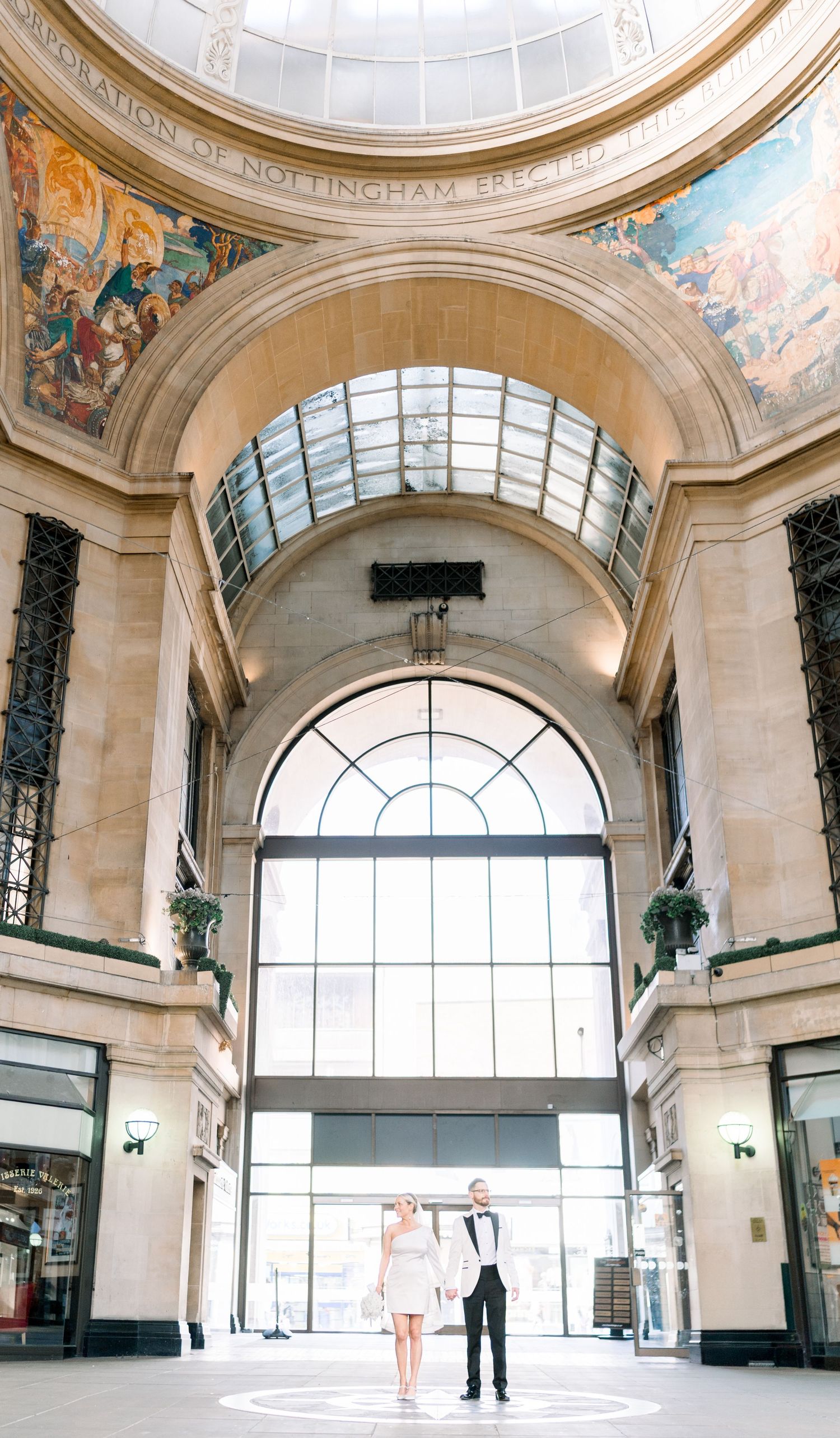 Bride & groom stand gazing up at grand cathedral-like architecture inside Nottingham Council House, with skylight, arches, and tall windows.