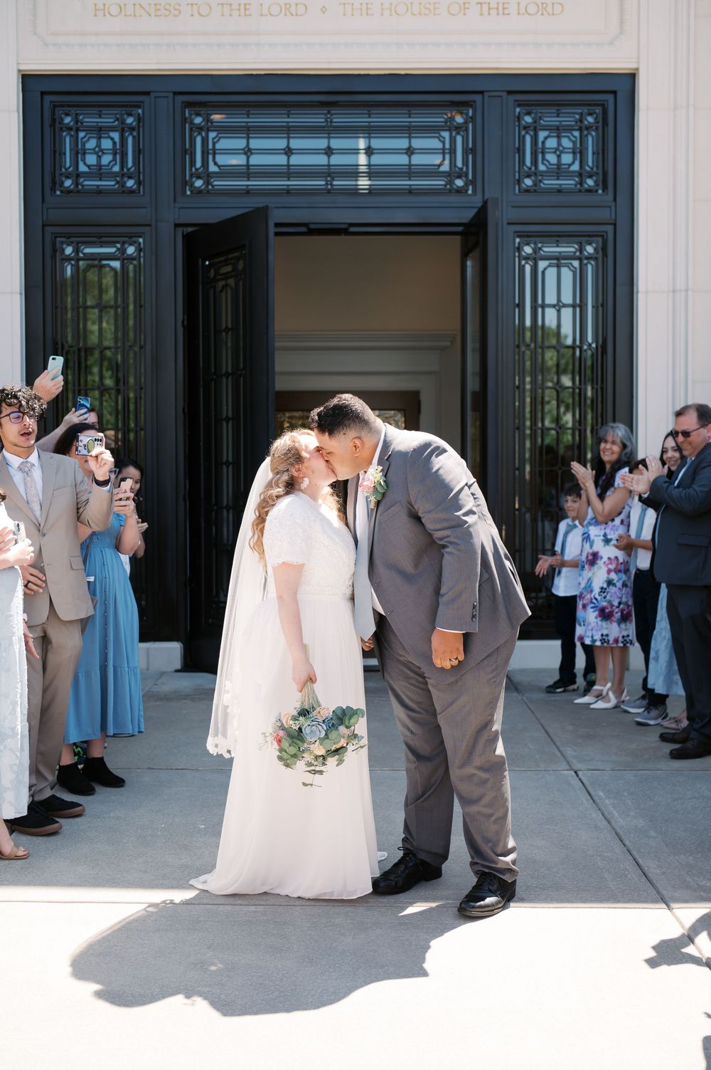 A couple shares a romantic kiss outside ornate church doors while wedding guests look on and celebrate the moment.