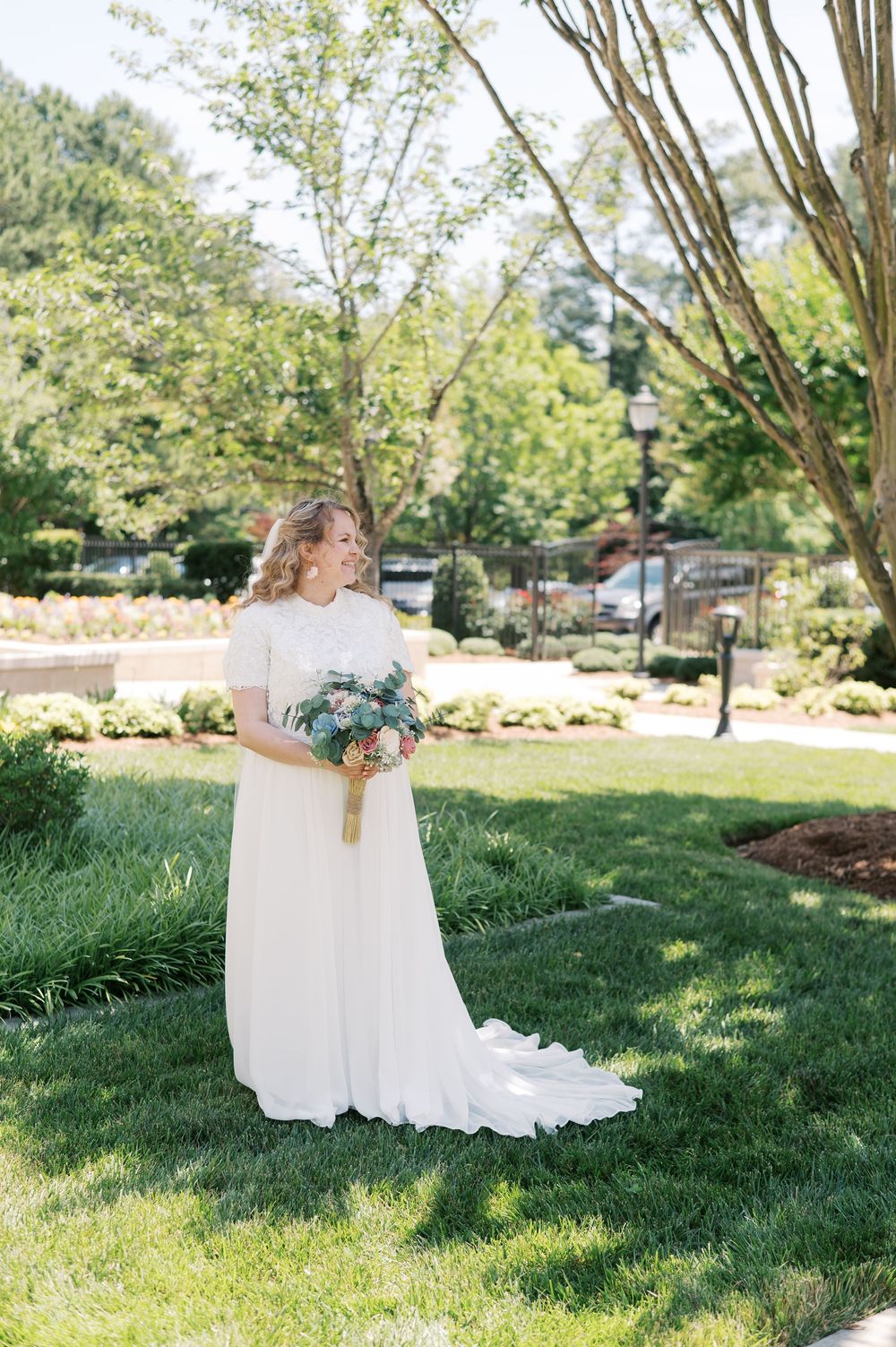 Someone in a flowing white wedding gown holds a blue and green bouquet while standing on manicured grass near trees and lamp posts.