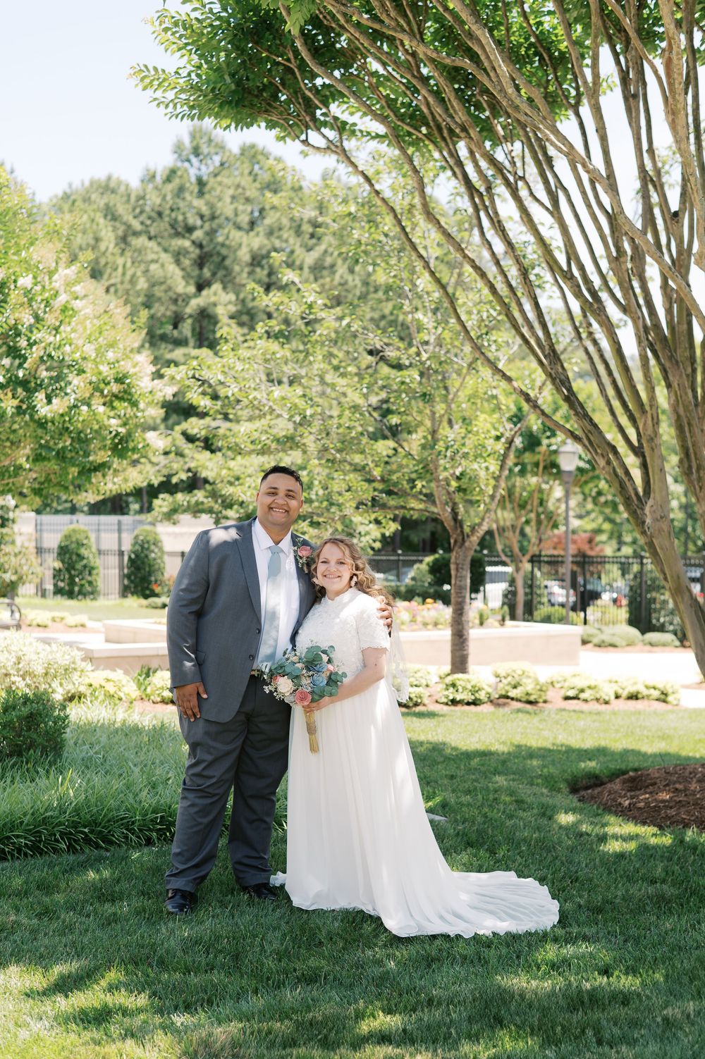 A wedding couple poses for a portrait on a beautiful green lawn with trees in the background.