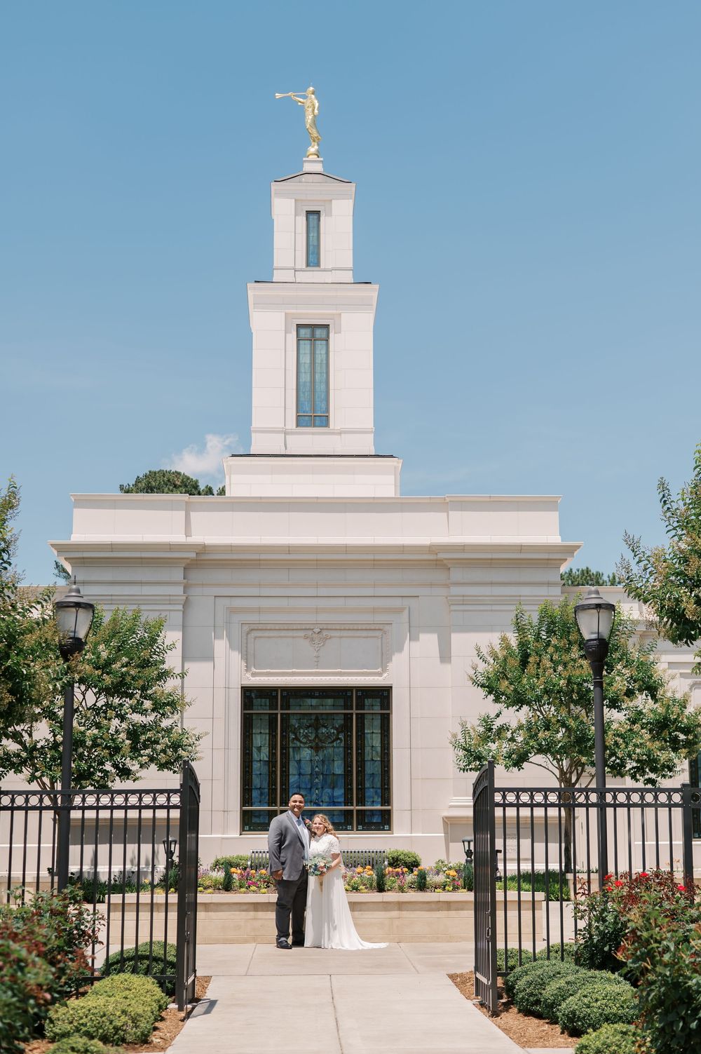 A white Mormon temple with a tall spire and golden angel statue stands behind a decorative iron gate with landscaped grounds.