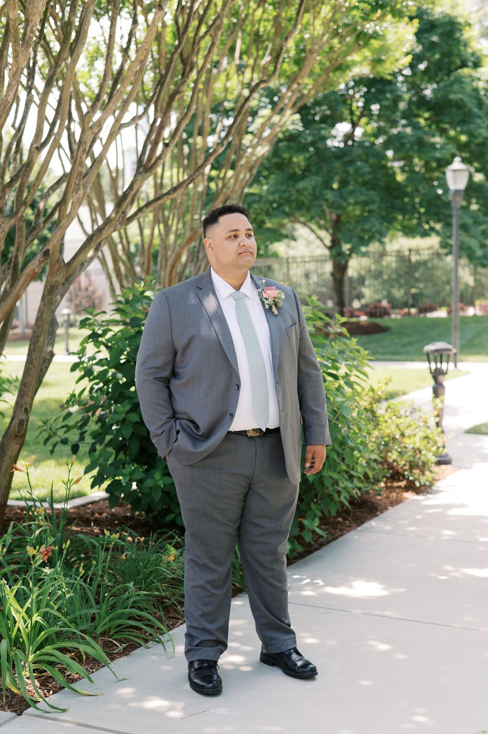 A person in a gray suit stands on a garden path under trees with a boutonniere pinned to their lapel.