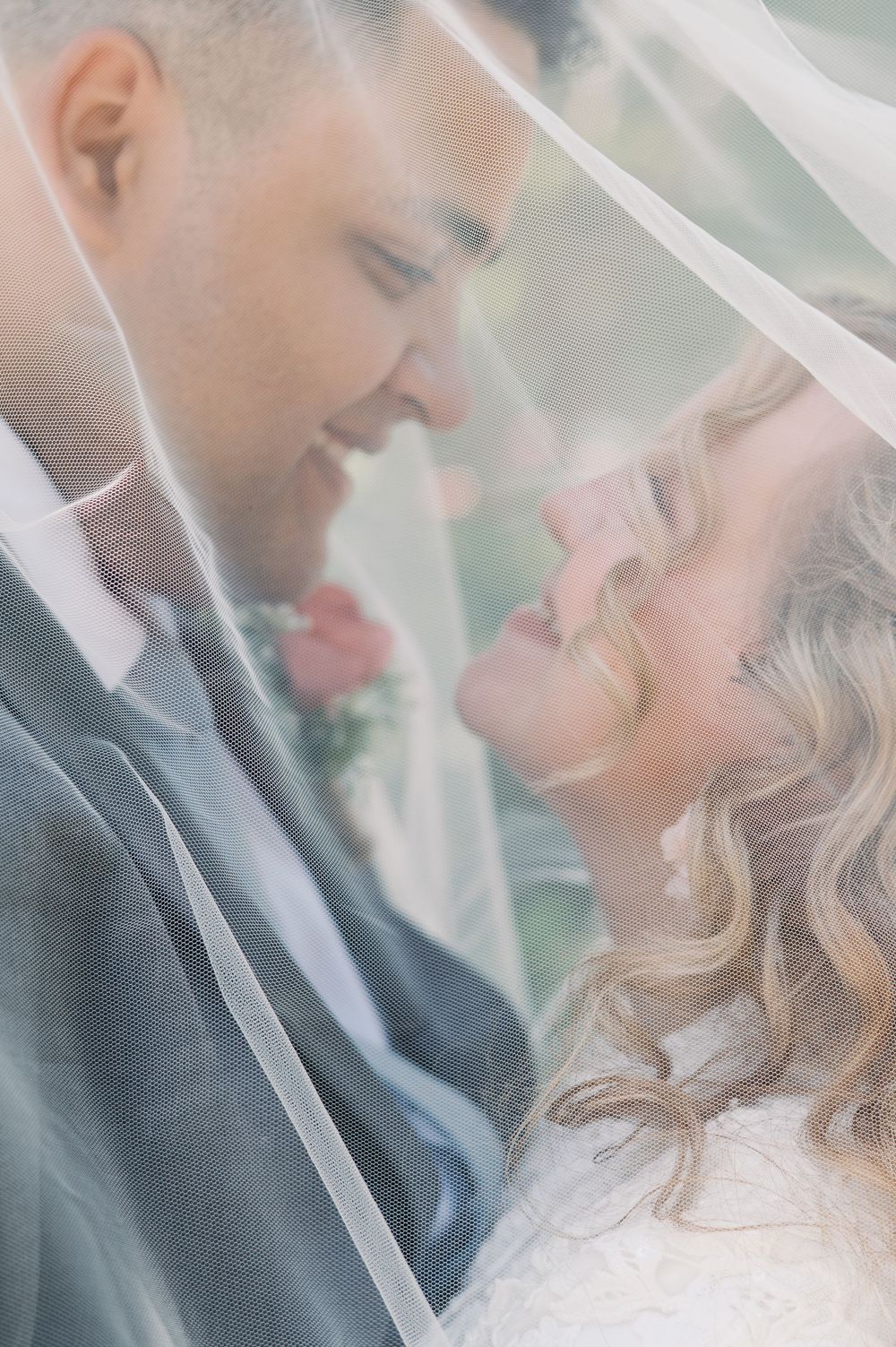 A romantic wedding couple shares an intimate moment under a flowing veil in soft dreamy light.