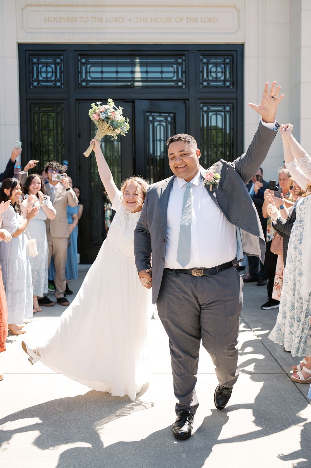 Joyful newlyweds exit their wedding ceremony while guests cheer and celebrate their union on a sunny day.