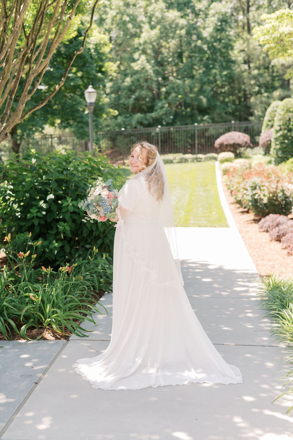 A person in a flowing white wedding gown and veil holds a bouquet while walking along a garden path on a sunny day.