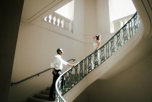 A figure in white ascends an elegant staircase with ornate mint green railings while natural light streams in from above.
