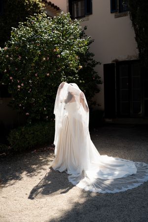 A flowing white wedding dress with a long train is displayed in a garden setting with dappled sunlight.