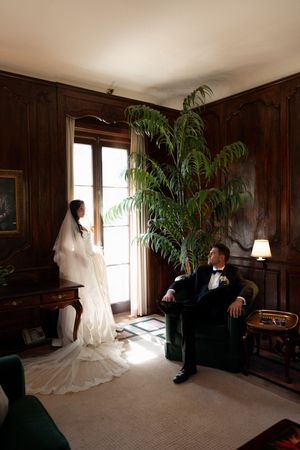 A couple in formal attire pose in an elegant wood-paneled room with a tall palm plant near French doors.