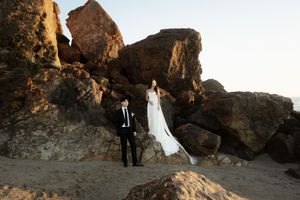 A couple in formal attire stands among dramatic desert rock formations at sunset for wedding photos.
