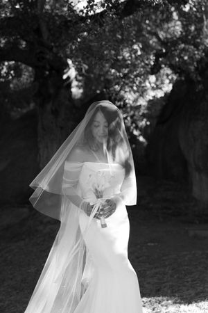Ethereal black and white bridal portrait showing bride in flowing off-shoulder gown and long veil standing in garden setting.