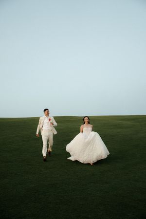 Two people in white formal attire joyfully running across a green lawn under an overcast sky.