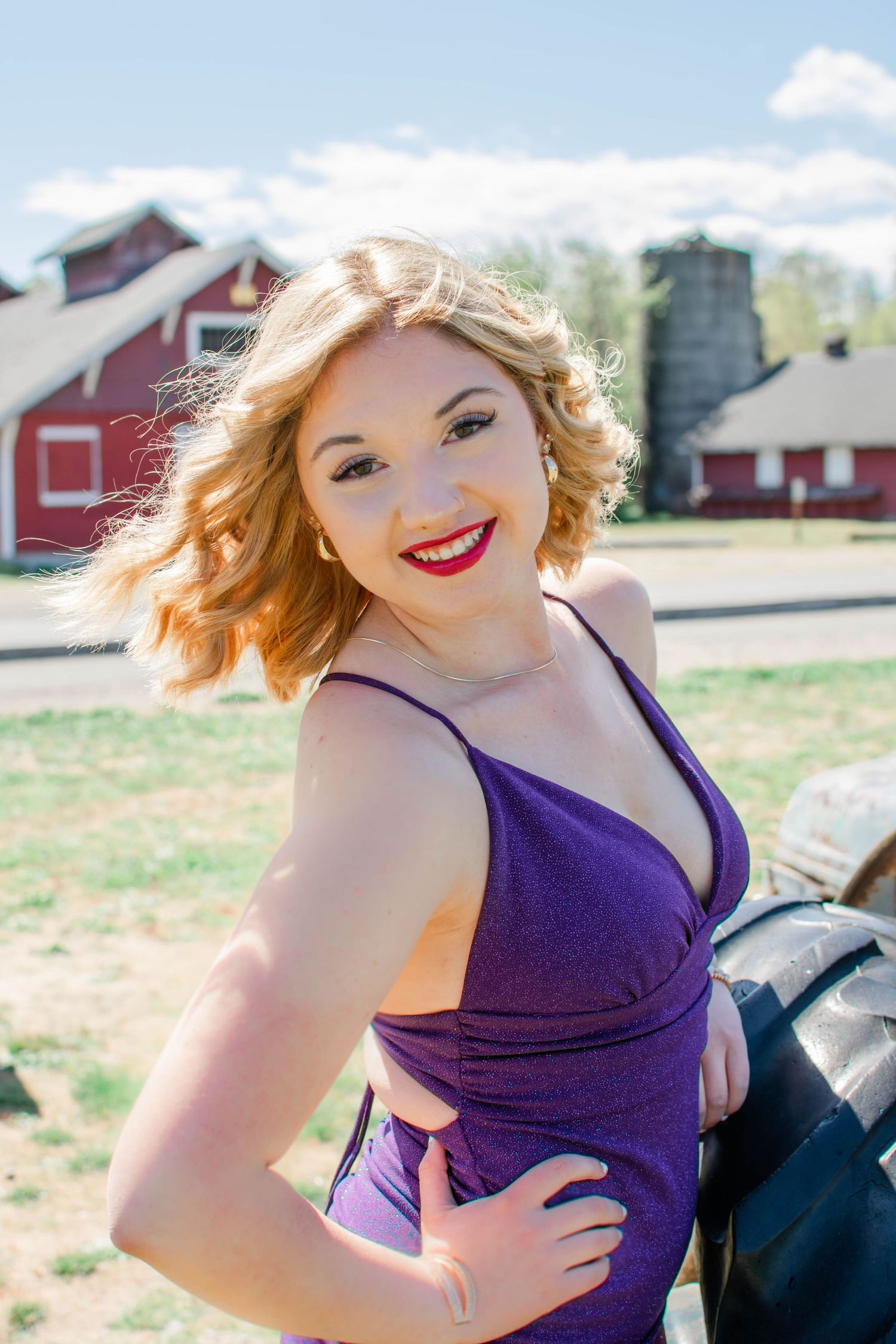 Graduate in purple dress poses by red barn on sunny day.